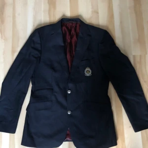 Morris marinblå kavaj med emblem - Klassisk marinblå kavaj från Morris med snyggt broderat emblem på bröstet och silvriga knappar med detalj. Fodret är vinrött och glansigt, vilket ger en lyxig känsla. Kavajen har flera fickor och en stilren passform, perfekt för dig som gillar preppy stil.