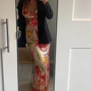 Blommig långklänning med cut-out - Säljer en snygg långklänning med blommigt mönster i rosa, rött, grönt och gult på ljus botten. Klänningen har en djup v-ringning, cut-out vid midjan och är ärmlös med smala axelband. Perfekt för sommarens alla tillfällen!