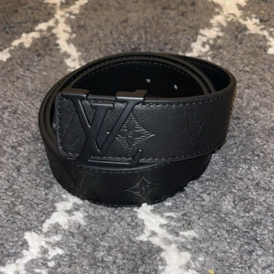 L-bälte  - L–bälte i svart med tonat monogram och svart spänne. Stilren och diskret design. Passar både herr och dam. Storlek 110 cm