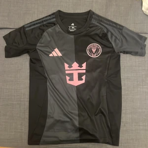 Inter Miami CF svart fotbollströja S - Svart och mörkgrå Inter Miami CF fotbollströja från Adidas i slim fit. Tröjan har korta ärmar, rosa broderade detaljer med klubbmärke och Adidas-logga samt ett stort rosa emblem på bröstet. Tillverkad i lätt och ventilerande polyester.