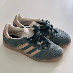 Adidas Gazelle sneakers i mocka - Adidas Gazelle sneakers i mörkgrön mocka med beige detaljer och klassiska tre ränder på sidan. Skorna har svart snörning, beige hälparti med Adidas-logga och en gummisula med mönster. Perfekt för dig som gillar retrostil och snygga färgkombos.