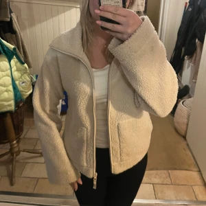 Beige teddyjacka från H&M - Mysig beige teddyjacka från H&M med stor krage och två rejäla fickor framtill. Jackan har en rak passform, lång ärm och stängs med dragkedja. Perfekt för dig som gillar chill och comfy stil under höst och vinter.