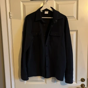 Svart overshirt från Selected Homme - Stilren svart overshirt från Selected Homme med klassisk krage och två bröstfickor med knappar. Perfekt lager-på-lager-plagg med lång ärm och snygga detaljer. Enkel att matcha och ger en clean look. Aldrig använd ny pris 800 mitt pris 500