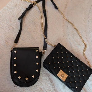 Två svarta axelväskor med gulddetaljer - Säljer två snygga svarta axelväskor i skinnimitation. Ena väskan har rundad form med guldnitar och justerbart axelband, den andra är rektangulär, quiltad med guldkedja och lås. Insidan har blommigt tyg. Perfekta för att lyfta din outfit!