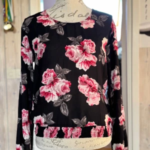 Blommig blus från H&M Divided - Säljer en svart blus från H&M Divided med stora rosa och vita blommor. Blusen har långa ärmar och resårdetalj nertill med blommönster. Perfekt för dig som gillar färgstarka prints och vill sticka ut lite extra.