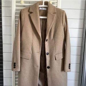 Beige kappa från Ellos med knappar - Stilren beige kappa från Ellos med klassisk krage, svarta knappar framtill och vid ärmslut. Jackan har två fickor med lock och är helfodrad. Perfekt för dig som vill ha en tidlös och snygg look under höst och vinter.