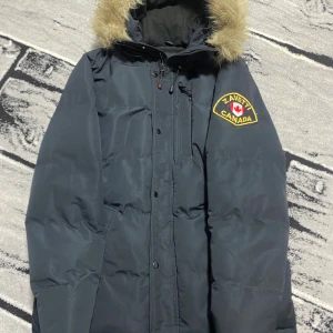 Svart dunjacka med päls från Zavetti Canada - Svart dunjacka från Zavetti Canada med snyggt broderat märke på ärmen och huva med fluffig päls. Jackan har dragkedja, flera fickor och en varm, quiltad insida. Perfekt för kalla vinterdagar och har en clean, urban look.