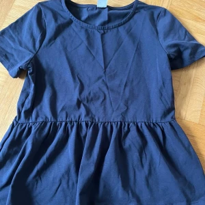 Mörkblå peplum topp i bomull - Säljer en mörkblå peplum topp med rund halsringning och korta ärmar. Toppen har en markerad midja med rynkad volang nedtill som ger en snygg siluett. Storlek 158/164