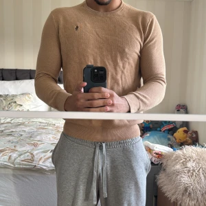 Beige stickad tröja från Polo Ralph Lauren - Snygg beige stickad tröja från Polo Ralph Lauren med den klassiska broderade loggan på bröstet. Tröjan har rund hals och långa ärmar, perfekt för dig som gillar stilren och enkel design. Passar dig som vill ha en clean look. Passar S med