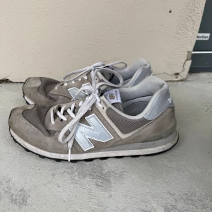 Grå New Balance 574 - Säljer ett par klassiska New Balance sneakers i grått med vita detaljer och stor N-logga på sidan. Skorna har snörning, platt sula och rund tå. Ovandelen är i mesh och mocka för skön komfort och snygg stil. Perfekta för dig som gillar retrovibe och streetwear. Välanvända med hål i hälkappan.