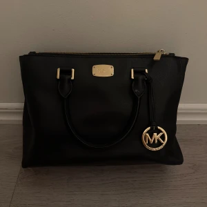 Svart handväska från Michael Kors - Stilren svart handväska från Michael Kors med guldfärgade detaljer och klassisk MK-logga. Väskan har två handtag, dragkedja upptill och är tillverkad i skinn. Insidan är fodrad med logotypmönstrat tyg. Perfekt för dig som gillar en lyxig och tidlös look.