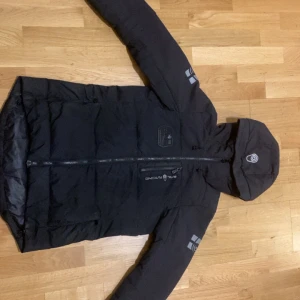 Svart dunjacka Sail Racing med huva - Svart dunjacka från Sail Racing med huva och reflekterande detaljer på ärmarna. Jackan har dragkedja framtill, flera fickor och snygga loggor på bröstet och huvan. Perfekt för kalla dagar och har en stilren, sportig look.