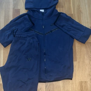 Blå Nike hoodie med dragkedja och byxor  - Säljer en marinblå hoodie från Nike med dragkedja och huva. Tröjan har korta ärmar och svarta detaljer över bröstet. Perfekt för dig som gillar sportig stil och vill ha något bekvämt och snyggt.