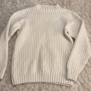 Vit ribbad stickad tröja - Säljer en vit stickad tröja med ribbad struktur och högre rund hals. Tröjan har långa ärmar och är perfekt för dig som gillar en enkel och clean look. Passar dig som vill ha något mjukt och skönt när det är kallt ute.