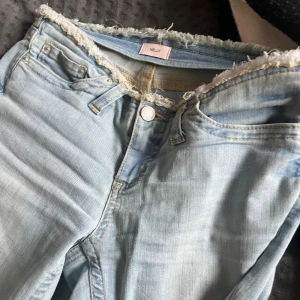 Ljusblå bootcut jeans från Nelly - Säljer ett par ljusblå jeans från Nelly med bootcut-modell och rå, fransad midja. Jeansen har klassisk femficksdesign och är tillverkade i mjukt denimtyg. Perfekta för dig som gillar en avslappnad och trendig look.