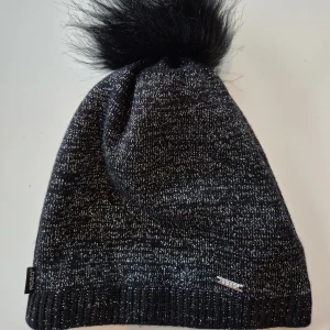 Snygg svart glittrig mössa med tofs  - Snygg svart glittrig mössa med tofs i fuskpäls. * 30% ull så denna håller dig varm :) * Onesize  Djur och rökfritt hem. Se bilder och förstora då de ingår i beskrivningen. Se gärna mina övriga annonser. Vid avhämtning finns den i Sollentuna.