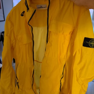 Gul vindjacka från Stone Island - Säljer en gul vindjacka från Stone Island med huva och svarta dragkedjor. Jackan har två bröstfickor med lock, två sidofickor med dragkedja och klassisk Stone Island patch på ärmen. Perfekt för dig som vill sticka ut med färg och stil.