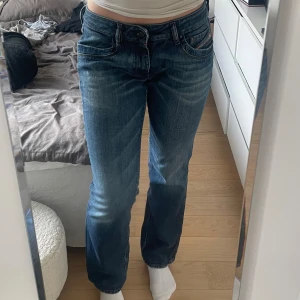 Diesel jeans lågmidjade - Säljer ett par blå lågmidjade jeans från Diesel, modell Ronhary. Jeansen har raka/utsvängda ben, och snygga slitningar framtill. De är tillverkade i stretchigt denim för extra komfort och har detaljerade fickor med Diesel-logga. Knappt använda, har satt i ett snöre i midjan för att spänna åt extra då de var för små på mig.