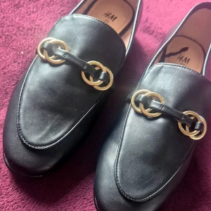 Svarta loafers med gulddetaljer H&M - Snygga svarta loafers från H&M i storlek 39. Skorna har en klassisk design med rund tå och guldfärgade ringdetaljer över foten. Tillverkade i skinnimitation med platt sula, perfekta för en stilren look. Endast testade. 
