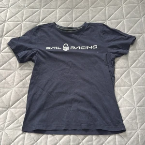 Mörkblå Sail Racing t-shirt - Mörkblå t-shirt från Sail Racing med vit logga tryckt över bröstet och en mindre logga i nacken. Klassisk rund halsringning med vit randig detalj på insidan. Skön och enkel modell i bomull, perfekt för en avslappnad stil.