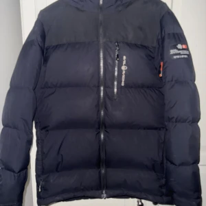Svart dunjacka Sail Racing Antarctica - Svart dunjacka från Sail Racing, modell Antarctica Expedition. Jackan har en puffig siluett, huva, flera dragkedjeförsedda fickor och snygga logodetaljer på bröstet och ärmen. Perfekt för kalla vinterdagar och riktigt stilren med sin mörka look.