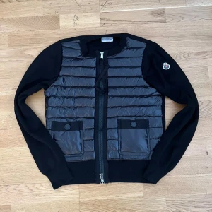 Moncler Cardigan  - Snygg svart cardigan jacka från Moncler med quiltad front i blankt material och stickade ärmar. Jackan har två fickor med knappstängning, dragkedja framtill och Moncler-logga på ärmen. Perfekt för dig som gillar stilren design och exklusiva märken.