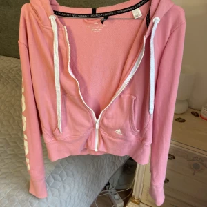 Rosa croppad hoodie från Adidas - Säljer en rosa croppad hoodie från Adidas med vit dragkedja, snörning och stor logga på ena ärmen. Tröjan har huva, kängurufickor och är tillverkad i bomullsmix med Climalite-teknologi. Perfekt för en sportig och chill look.storlek passar 34-38