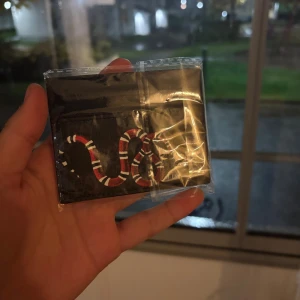  svart korthållare med orm - Snygg svart korthållare från i skinn med klassiskt GG-mönster och en cool röd, vit och svart orm på framsidan. Perfekt för dig som vill ha något unikt och stilrent i fickan. Flera kortfack på båda sidor.