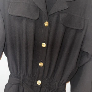 Svart jumpsuit från Saint Germain - Svart jumpsuit från Saint Germain med guldknappar framtill, krage och två bröstfickor. Jumpsuit har  markerad midja med resår och knytskärp. Knappt använd