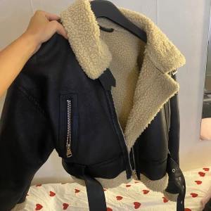 Svart pälskantad bikerjacka med bälte - Snygg svart bikerjacka med beige fuskpälsfoder och krage. Jackan har dragkedjor, bälte i midjan och detaljer i silver. Perfekt för dig som vill ha en cool och varm look under höst och vinter. Helt ny använd en gång jätte bra skick 