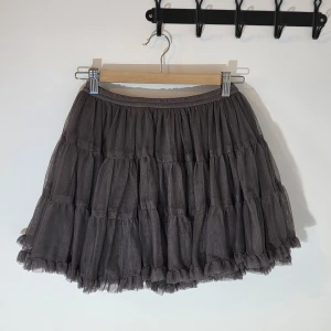 Mörkgrå volangkjol från Mothercare - Supersöt mörkgrå kortkjol från Mothercare med flera lager av mjuk tyll och volanger. Kjolen har en elastisk midja och är fodrad med bomull för extra komfort. Perfekt för dig som gillar fluffiga och lekfulla kjolar med mycket rörelse.