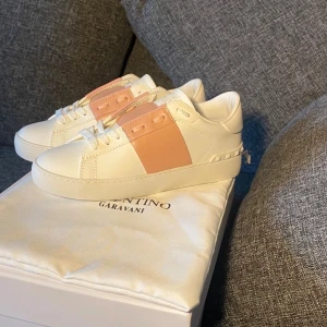 Valentino Garavani Open sneakers vit/rosa - Snygga Valentino Garavani Open sneakers i vitt skinn med bred ljusrosa läderdetalj över snörningen. Platt sula med mönstrad undersida och rund tå. Ikonisk design med subtila nitar på hälen och vita snören. Perfekt för dig som gillar exklusiva sneakers med stil. Kvitto och äktenhetsbevis finns!