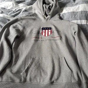 Grå hoodie från GANT med broderad logga - Säljer en grå hoodie från GANT i storlek XL. Tröjan har broderad logga och sköld på bröstet, känguruficka och justerbar huva med snörning. Tillverkad i mjuk bomullsmix, perfekt för chill dagar.