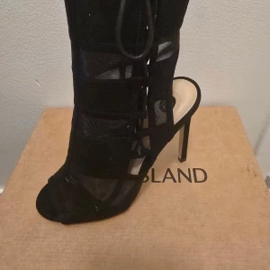 Svarta höga klackskor från River Island - Säljer ett par svarta klackskor från River Island med öppen tå och snörning framtill. Skorna har höga smala klackar och är tillverkade i en kombination av mesh och mockaliknande material. Snyggt statement med coola detaljer och trendig design.