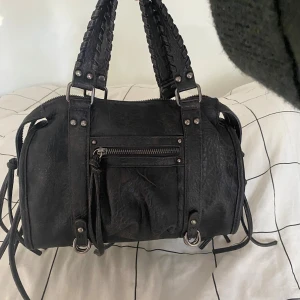 Svart axelväska med flätade handtag - Cool svart axelväska i skinnimitation med flätade handtag och justerbar axelrem. Väskan har silverfärgade metalldetaljer, dragkedja framtill och snygga knytband på sidorna. Perfekt storlek för att få plats med allt du behöver till vardagen.