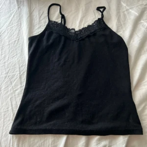 Svart linnetopp med spetskant - Snygg svart linnetopp med smala axelband och dekorativ spets längs urringningen. Perfekt att styla lager på lager eller bära som den är. Enkel och stilren design som passar till många olika looks.