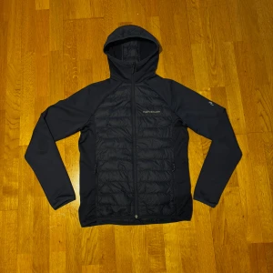 Peak Performance Hybrid Jacka - Snygg svart pufferjacka från Peak Performance i storlek M. Jackan har huva, hel dragkedja och quiltad framsida med broderad logga på bröstet. Materialet är polyester och syntet, perfekt för kyliga dagar. Stilren design med långärmade ärmar och två sidofickor med dragkedja. Helt ny, använd fåtal gånger. Kan gå ner i pris vid snabb affär. Om frågor komm pv!