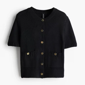 Svart stickad kofta med guldknappar - Kort svart stickad kofta med guldiga knappar framtill och två fickor. Koftan har korta ärmar och en ribbad kant vid halsen och ärmsluten. Perfekt att slänga över ett linne för en chill och snygg look.