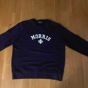 Mörkblå sweatshirt från Morris - Snygg mörkblå sweatshirt från Morris med vit broderad logga och emblem på bröstet. Tröjan har rund hals, ribbade muddar och lång ärm. Tröjan är i storlek L fast krympt så den passar folk med storlek M även S. Tröjan är inte förstörd nånstans utan hel på alla plan.