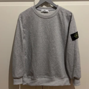 Grå sweatshirt från Stone Island size S - Snygg grå sweatshirt från Stone Island med klassisk rund hals och ribbade muddar. Tröjan är i nytt skick och har den ikoniska svarta patchen med logga på vänster ärm. Perfekt för dig som gillar stilrena och sportiga plagg. Size S