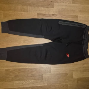 Svarta Nike mjukisbyxor med dragkedja - Svarta mjukisbyxor från Nike med gråa paneler längs benen och röd Nike-logga på framsidan. Byxorna har snörning i midjan, ribbade muddar och en dragkedjeficka på höger sida. Perfekta för chill eller träning.