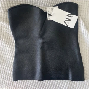 Svart tubtopp från Zara - Snygg svart tubtopp från Zara med stilren bandeau-design. Toppen är axelbandslös och har en slät, elegant yta som ger en cool och modern look. Perfekt att styla med jeans eller kjol för en trendig outfit.