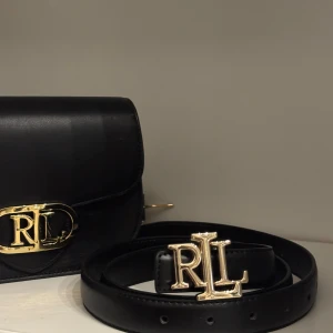 Svart RL bälte med guldfärgat spänne - Snyggt svart bälte från Ralph Lauren i slätt skinn med ett stort guldfärgat RL-spänne framtill.110Cm 