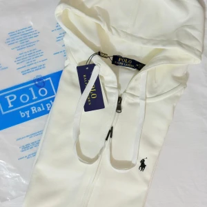 Vit hoodie från Polo Ralph Lauren - Snygg vit hoodie från Polo Ralph Lauren med klassisk svart logobrodyr på bröstet. Tröjan har huva med snörning och dragkedja framtill. Tillverkad i mjuk bomull som känns skön mot huden. Perfekt för chill dagar och streetwear-stil.