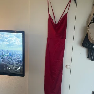 Röd långklänning med snörning - Säljer en snygg röd långklänning med tunna axelband och djup rygg med snörningsdetalj. Klänningen har en slimmad passform och är gjord i ett glansigt satintyg som ger en lyxig känsla. Perfekt för dig som vill sticka ut med en elegant look.