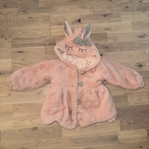 Rosa teddyjacka med kaninöron - Söt rosa jacka med kaninöron på huvan och en grå rosett. Jackan har broderade ögonfransar på huvan och stängs med tre knappar. Den har en lätt utställd modell. Supersöt rosa teddyjacka med huva som har broderade ögon, grå rosett och kaninöron. Jackan är fluffig och har knappar framtill samt ett glansigt innerfoder. Perfekt för dig som vill ha en gullig och mysig look. Liten fläck går säkert att tvättas bort annars är den är mycket bra skick👌🏻 från cirka 1år + 
