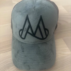 Grå Maggiore keps i mocka och mesh - Snygg grå keps från Maggiore med mockafront och svart mesh på baksidan. Framtill finns en stor svart broderad logga. Kepsen har böjd skärm och justerbar passform. Perfekt för dig som vill ha en cool och modern look.