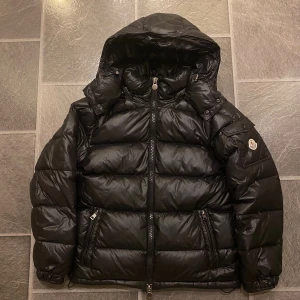 Svart Moncler pufferjacka med huva - Säljer en svart pufferjacka från Moncler med huva och dragkedja framtill. Jackan har två fickor med dragkedja, snygg design och klassisk Moncler-logga på ärmen. NFC fungerar och sitter i Moncler-loggan. Den är köpt december 2024. Jag har kvittot online. Har kvar en hängetikett som kom med jackan. Priset kan diskuteras!