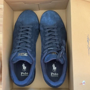 Mörkblå sneakers från Polo Ralph Lauren - Snygga mörkblå sneakers från Polo Ralph Lauren med mockayta och klassisk snörning. Skorna har rund tå, platt sula och guldfärgad Polo-logga på hälen. Perfekt för dig som gillar stilrena och tidlösa sneakers med exklusiv känsla.