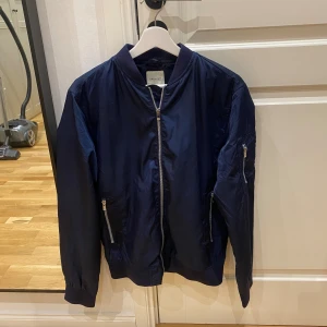 Mörkblå bomberjacka Jack & Jones  - Snygg mörkblå bomberjacka från Jack & Jones Premium. Jackan har klassisk bomberkrage, silverfärgade dragkedjor på fickor och ärm, samt ribbade muddar. Tillverkad i lätt och glansig polyester, perfekt för dig som vill ha en stilren och enkel look.